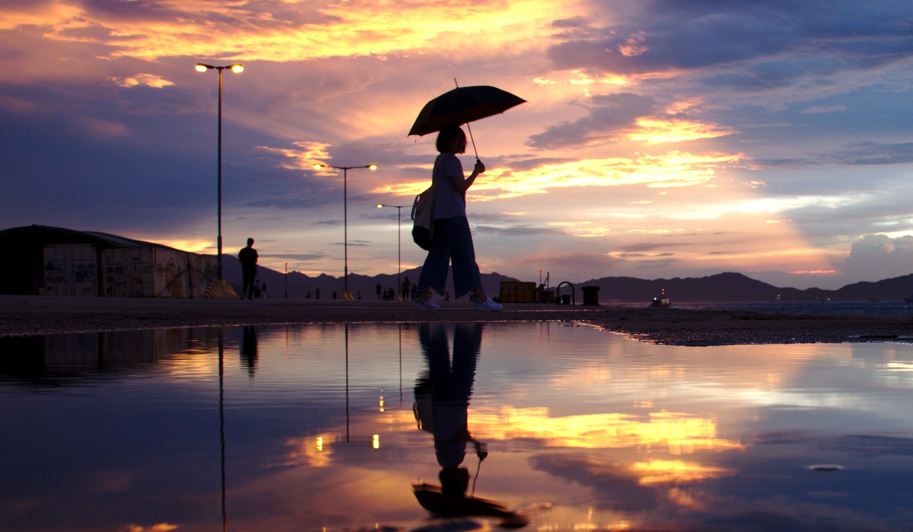 A backlit figure stands holding an umbrella at sunset. The yellows and blues of the sky are reflected in the ground which is wet after rain.