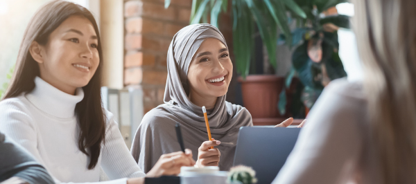 two women seated at a business meeting table smile at a woman across from them.