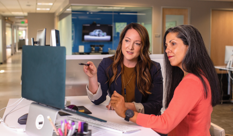 Two women in professional attire sit together at a desk discussing items on a desktop computer monitor.