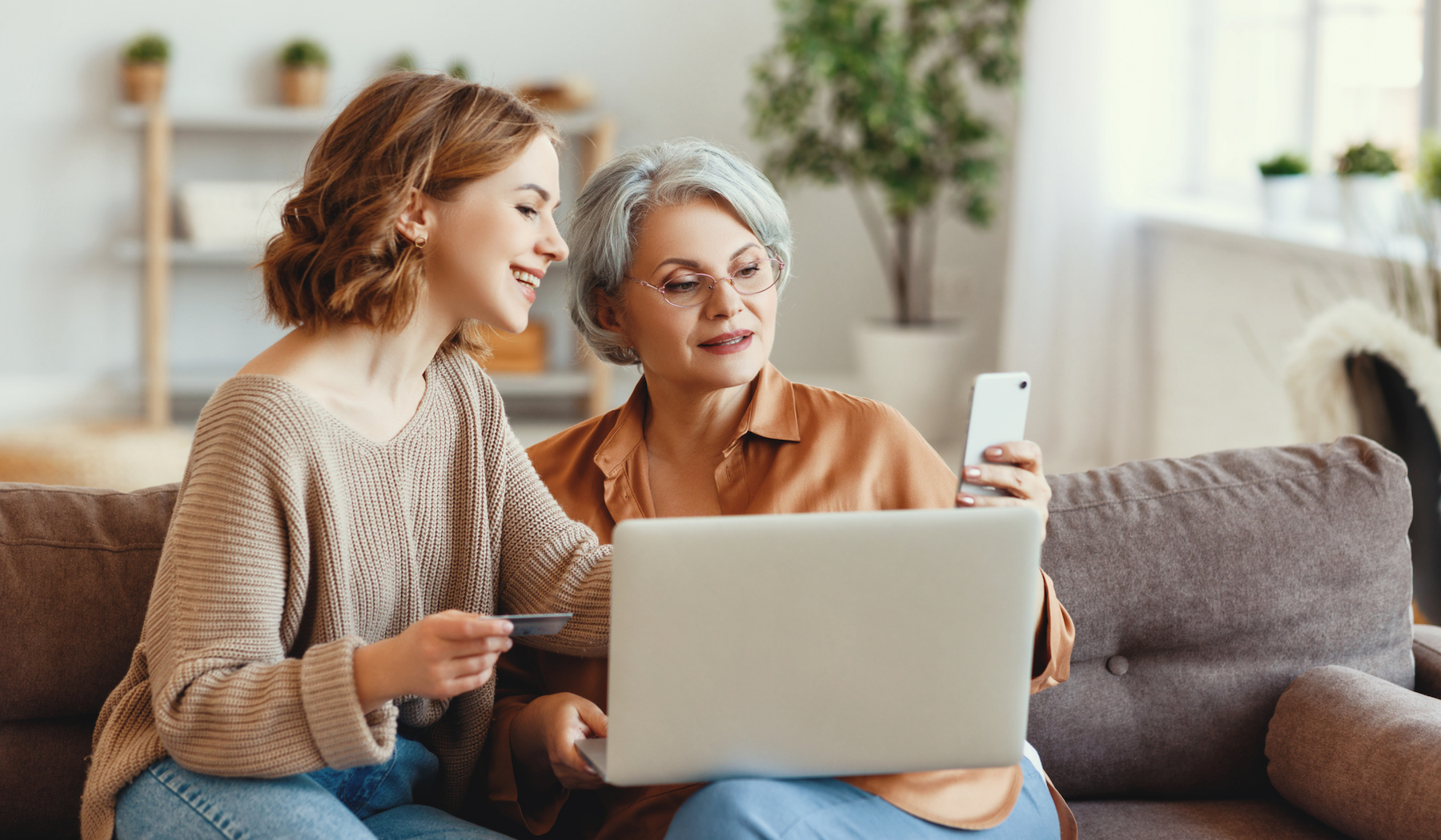 A mother and daughter sit on a couch together looking at a laptop screen. The mother is holding a cellphone while the daughter holds a credit card.